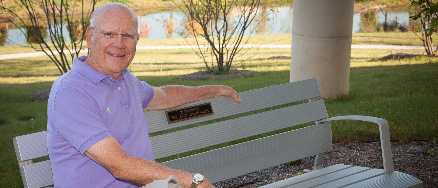 一名男子坐在长凳上，上面写着“纪念黛安·诺里斯博士”.D., RN"