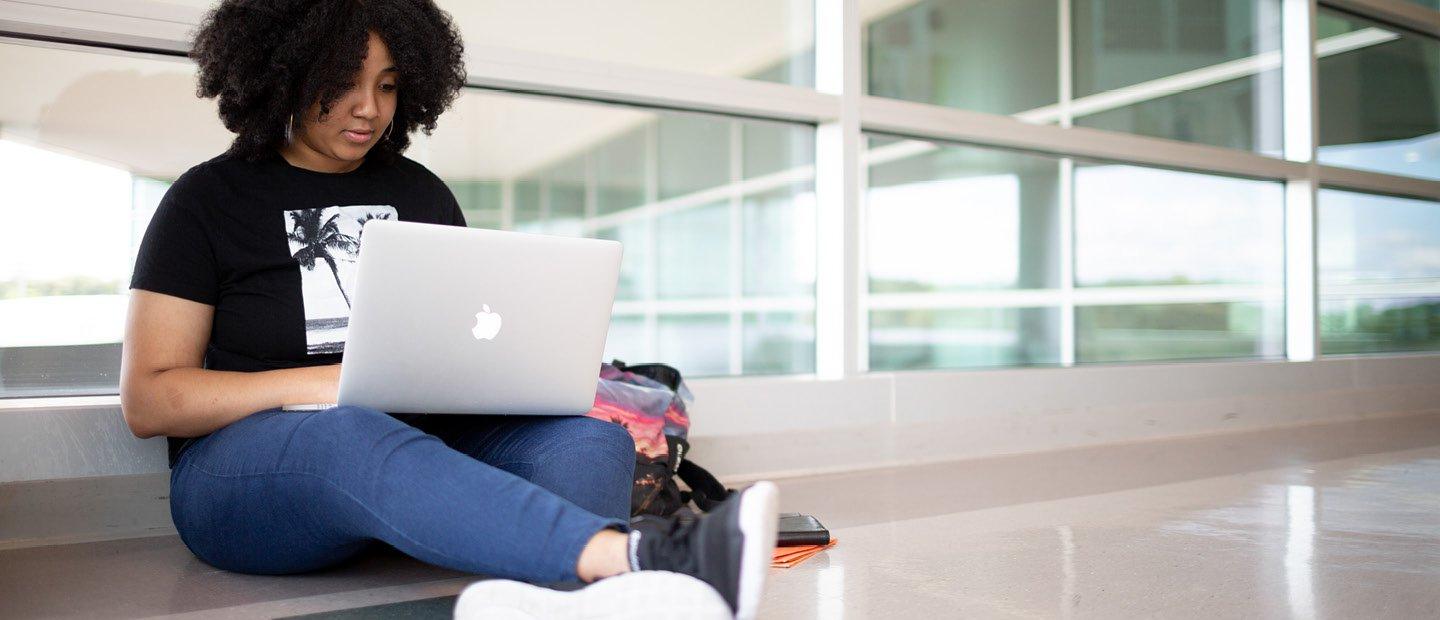 一位年轻女子坐在窗前的地板上，膝上放着一台打开的笔记本电脑