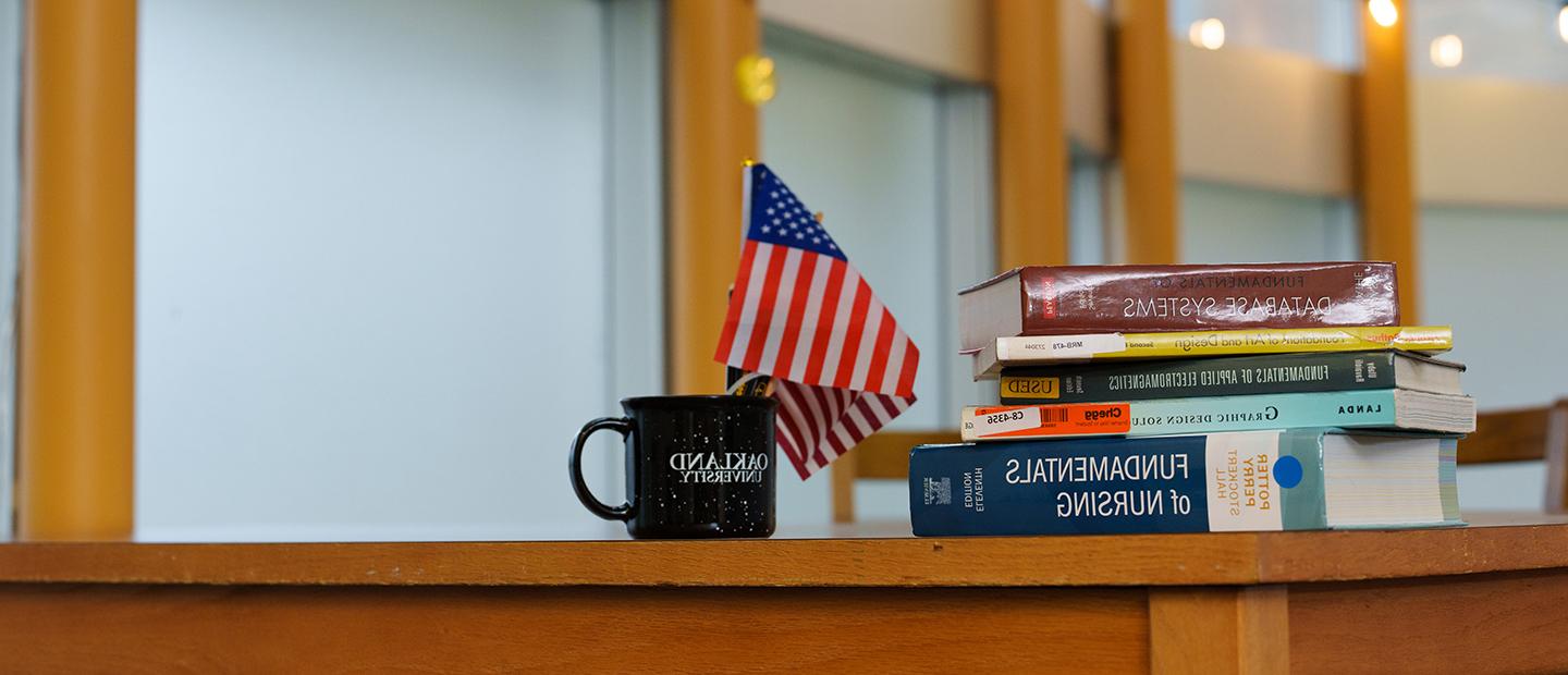 桌子上堆满了书，旁边是一面美国国旗和一个咖啡杯