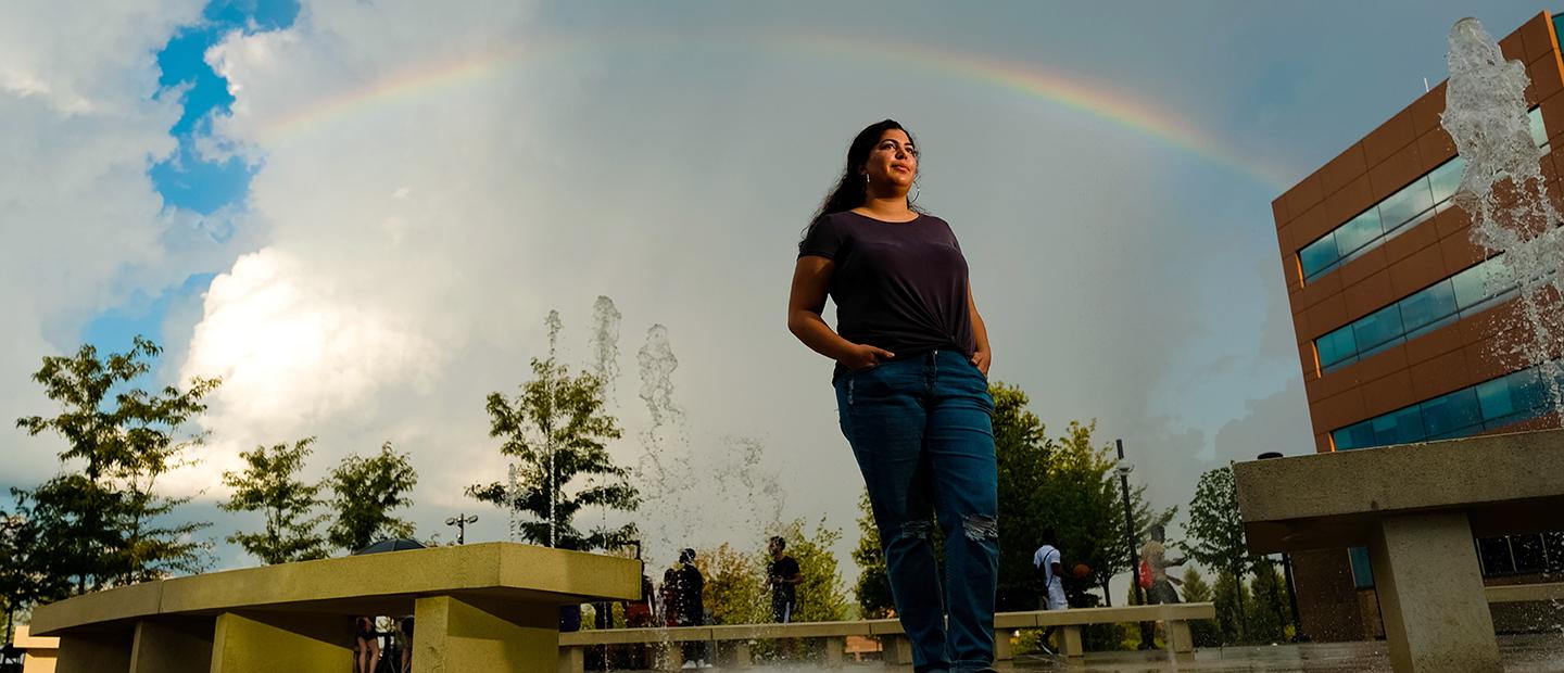 一位年轻女子站在韦德体育app官网的校园里，双手插在口袋里, 身后有彩虹和喷泉.