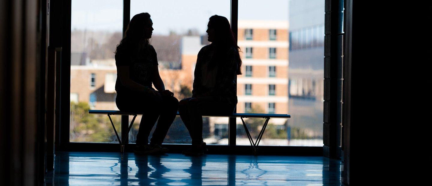 Silhouettes of two people seated on a bench in front of a window with O U campus in the background